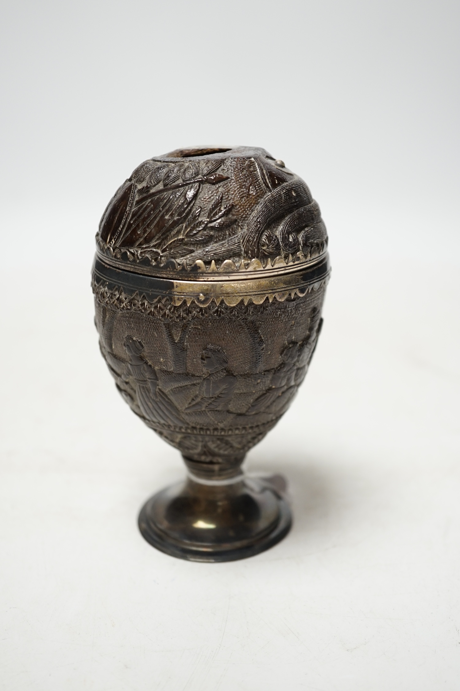 An early 19th century white metal mounted coconut cup, carved with continuous scene of figures, height 13.8cm. Condition - poor